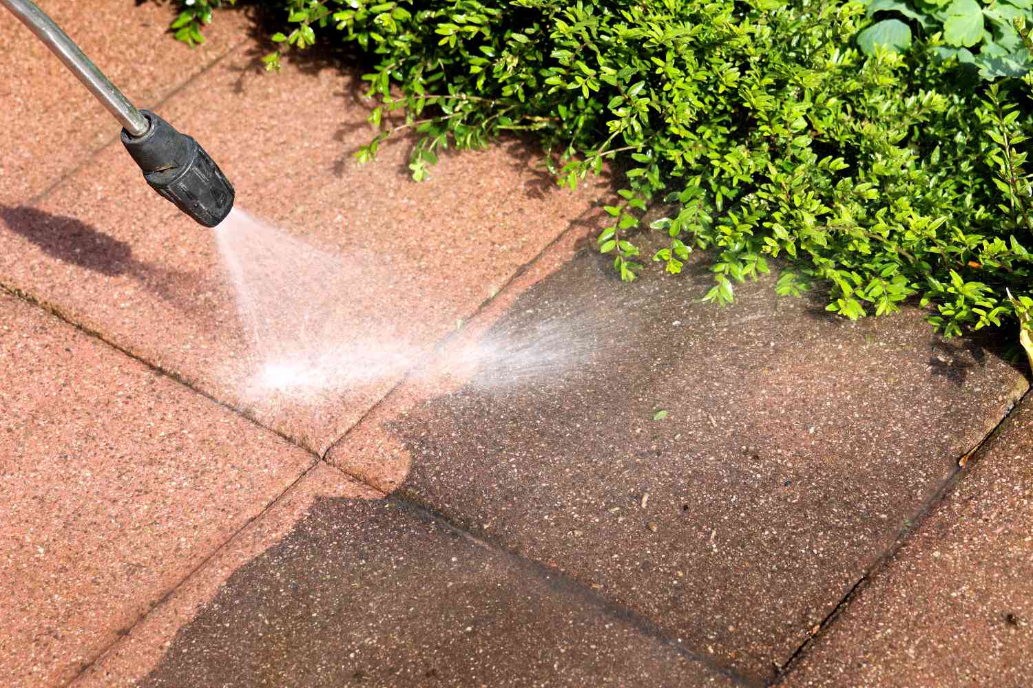 Pressure washer cleaning dirty pavers beside a hedge